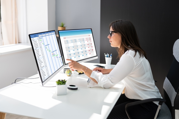 Side profile of a white woman with shoulder length dark hair, wearing a white shirt. She is sat at a desk in front of two monitors. The left hand monitor displays a Gantt chart, the right hand monitor shows a calendar. The walls are white and black.