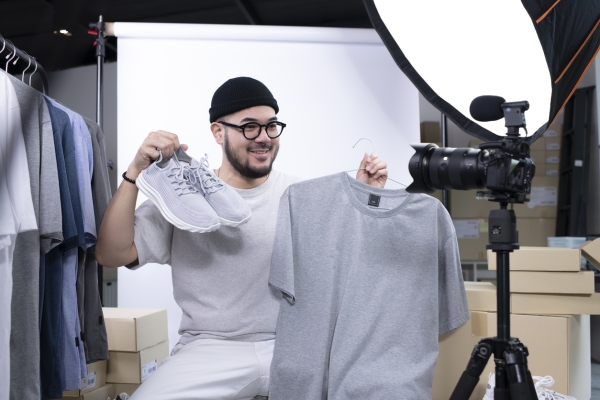 A male fashion influencer holds a pair of grey trainers and grey t-shirt while sat looking towards a camera. Around him are a circular studio light, a paper screen, and a selection of t-shirts hanging up on a rail