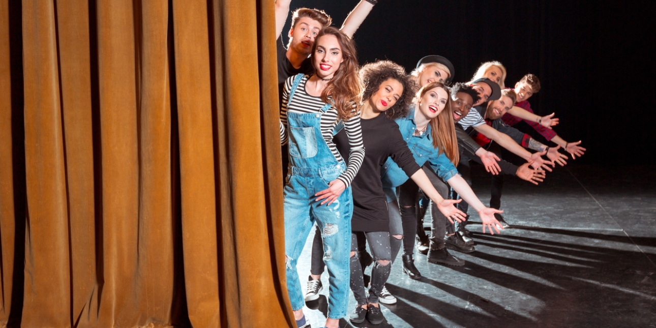 Ten young adult actors of different genders and ethnicities stand in a line on stage to the right of a theatre curtain. Most have their arms extended outwards in a jazz hands formation.