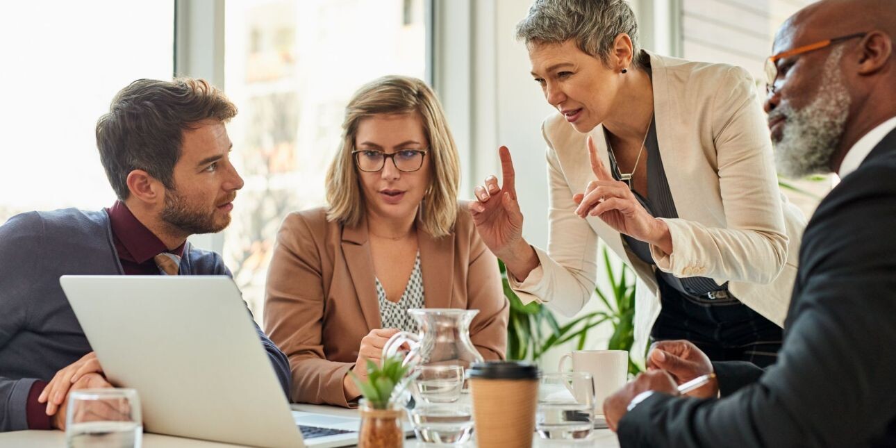 Huddled around a laptop, a white woman with short grey hair stands with her fingers raised while three colleagues listen - a younger white man with brown hair, a younger white woman and a Black man with grey beard - look on.