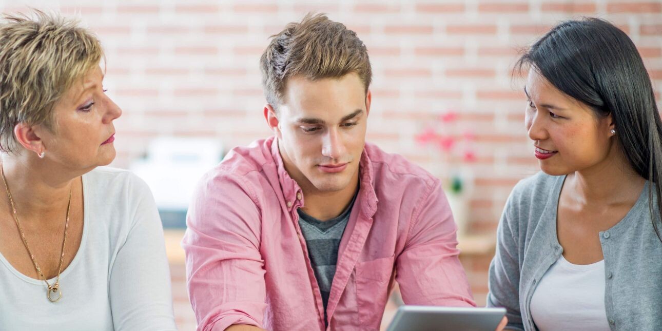 A young white man with short blond hair and pink shirt stares at a tablet. He is flanked on his left by an Asian woman with long dark hair and grey cardigan. On his right is an older white woman with short blonde hair and white top.