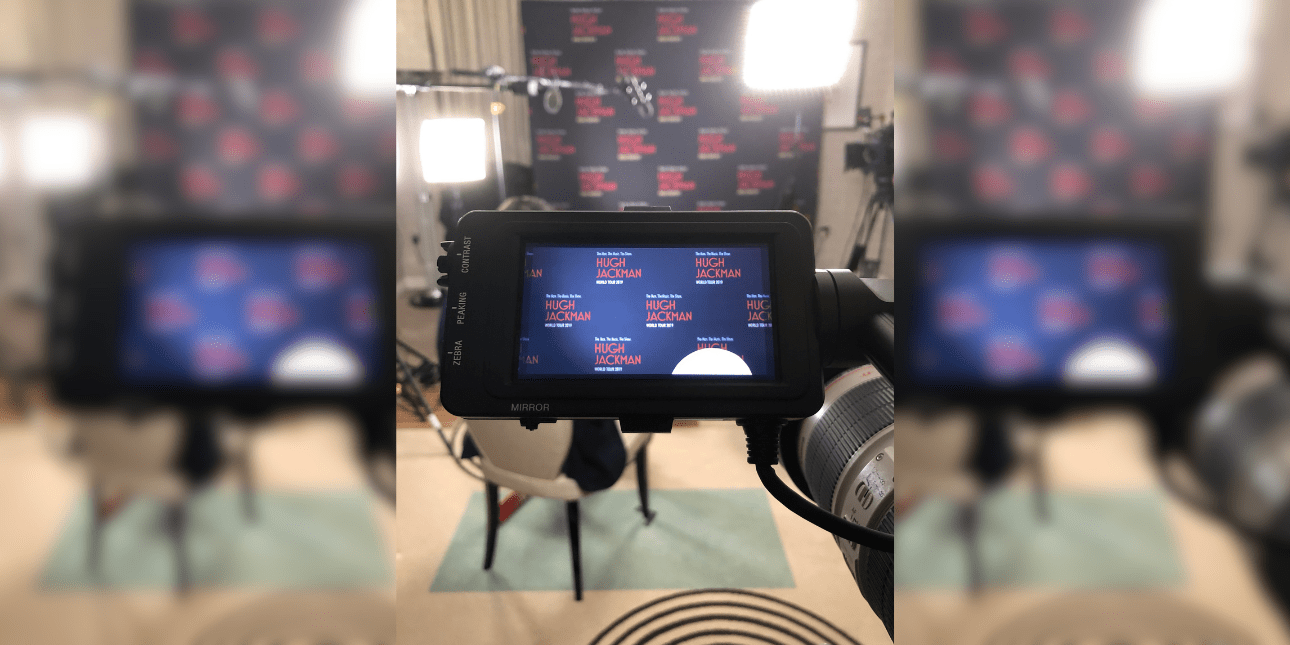 Looking through a video camera screen at the background of an empty chair, portable lights and boom microphone which are situated in front of a dark blue screen with red type which reads Hugh Jackman world tour