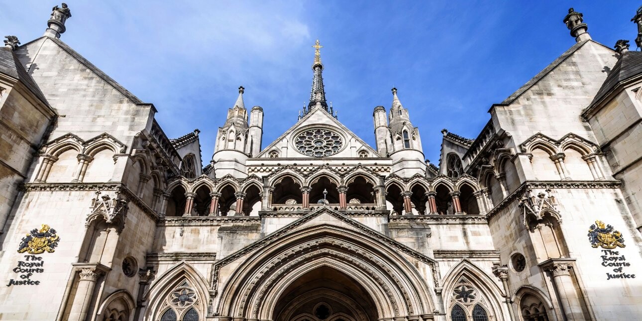 The symmetrical stone facade of the Royal Courts of Justice featuring an arched central section, towers, turrets and a gable containing a rose window above the entrance. A crest and the building name sit either side of the entrance.