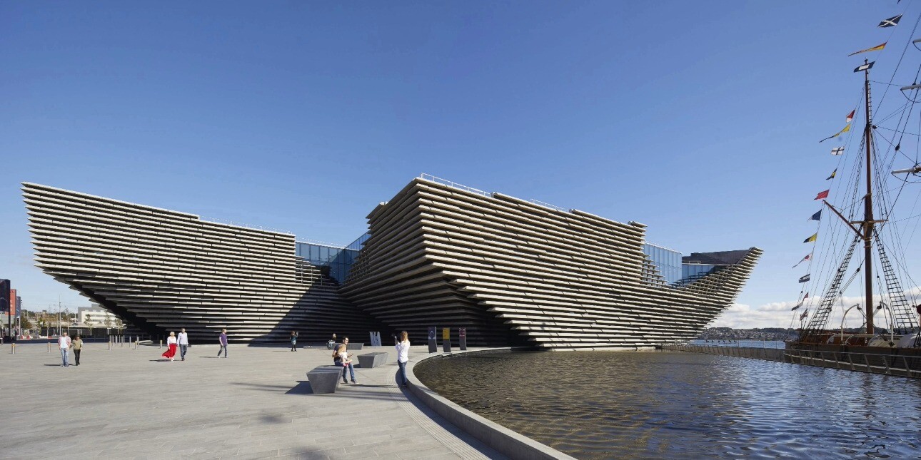 The exterior of the V&A Dundee cladded with lines of pre-cast reconstituted stone panels that run horizontally around curving concrete walls. The shape takes inspiration from cliffs with the prow jutting over the River Tay like a boat. The sky is bri