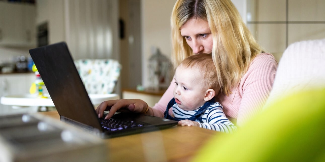 At a kitchen table a white woman with blonde hair kisses her baby's head sat in her lap while working at her laptop
