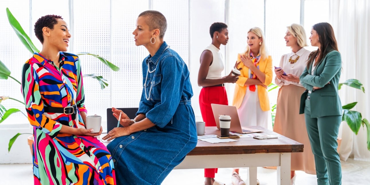 Six business women of different ages and ethnicities. Two in the foreground are perched on a table. The others are stood chatting behind. All are wearing bright and colourful clothes.