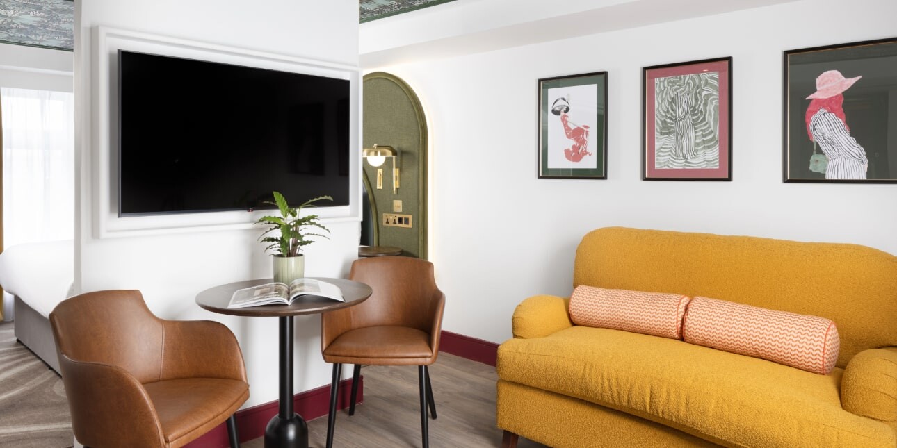 A picture of a hotel suite with white walls with pictures on them and a wall mounted TV, a table and brown chair and a yellow sofa
