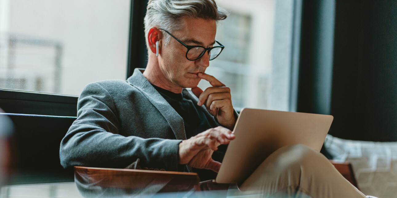 A white man with grey hair, wearing black-rimmed glasses, a grey jacket and a white headphone in his ear, stares at a laptop rested on his lap. He rests one finger on his chin and another on the laptop. There is a window in the background