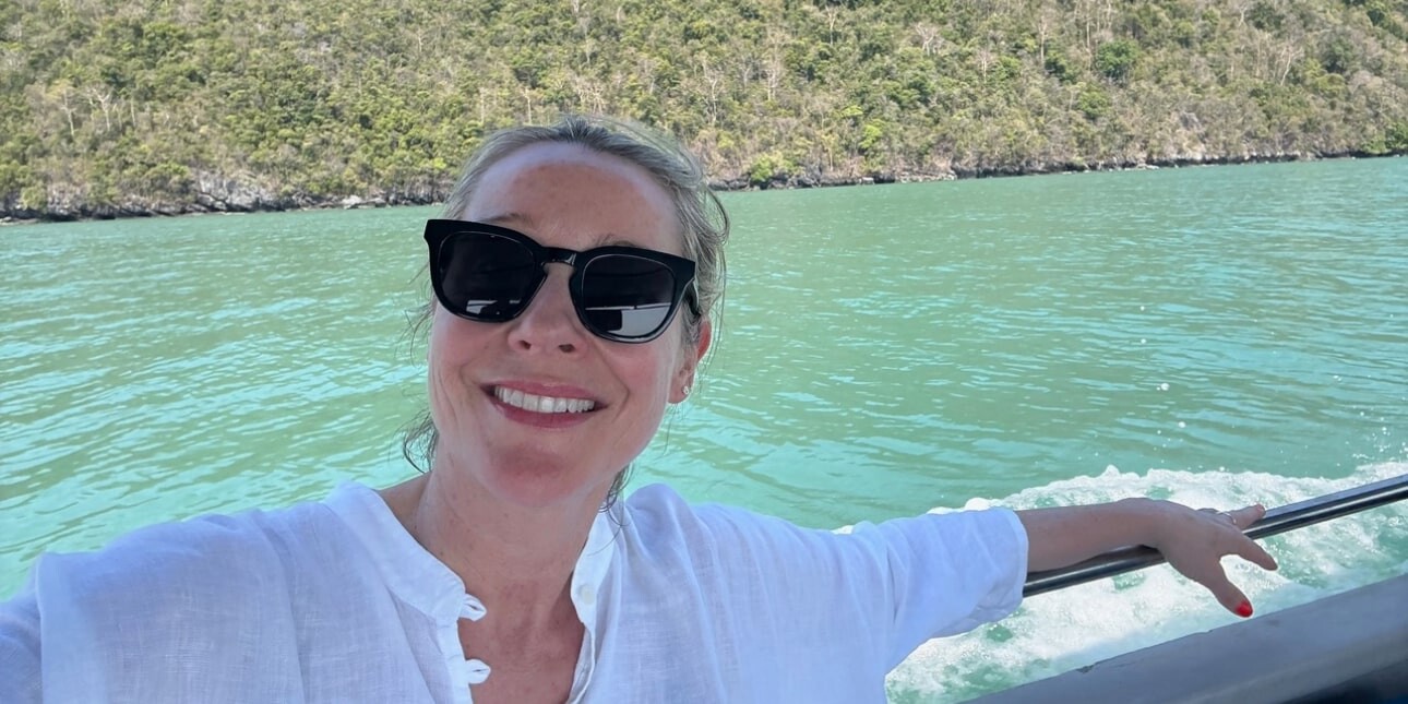 Juliet Kinsman is a blonde white woman wearing dark glasses. She wears a white shirt and is sat with her hand over a rail with an expanse of water and the greenery of the shore behind her.