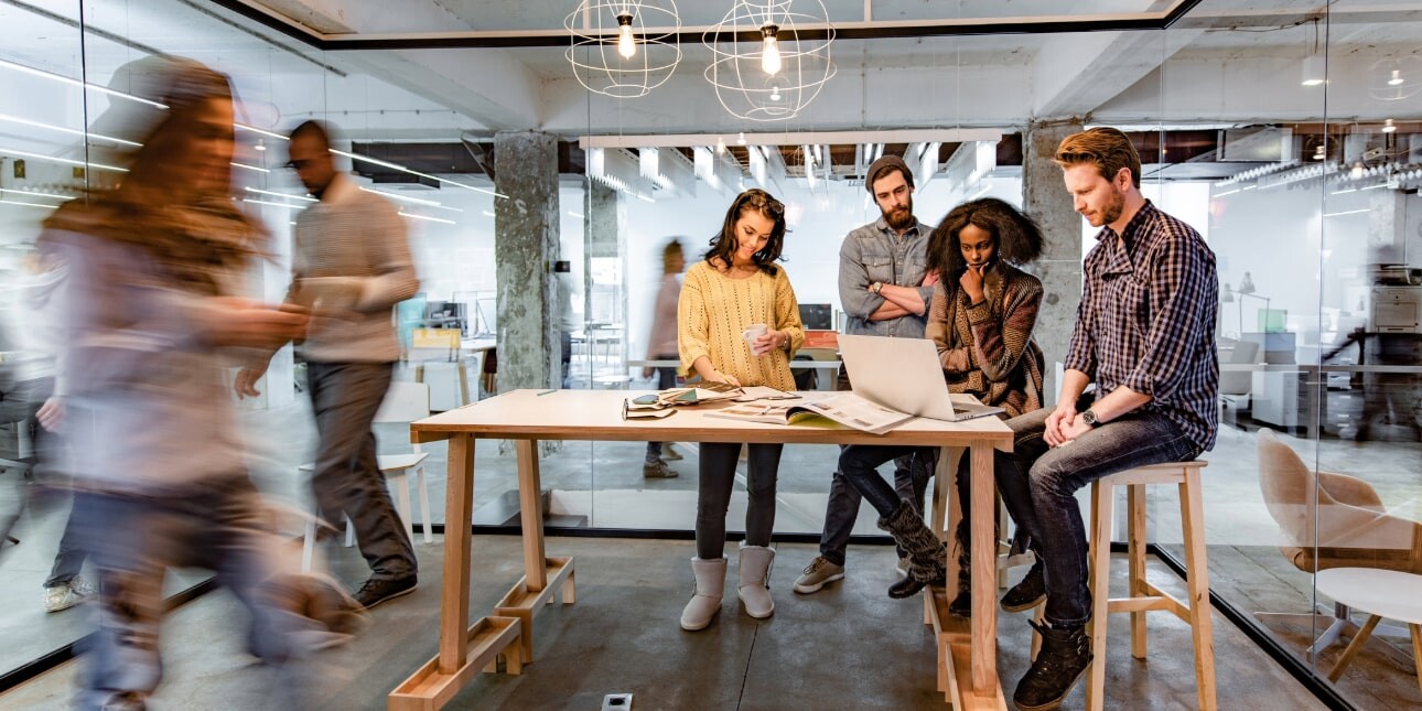 Two white men, a black woman and a white woman sit around a table looking at a laptop while other figures are blurred in the bacground walking past the glass office in which they are sat or stood