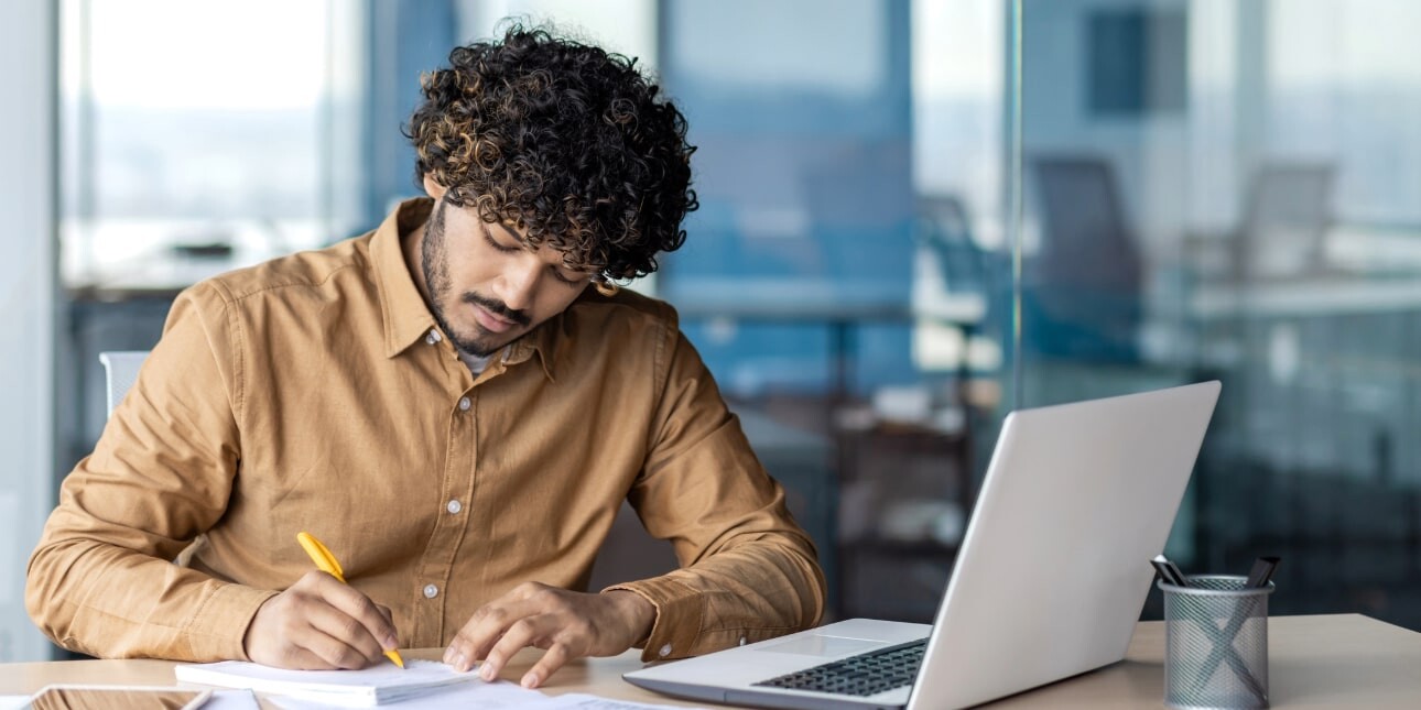 An Asian man with curly dark hair, who is wearing a mustard coloured shirt, writes on a notepad. He is in an office and there is a laptop open in front of him.
