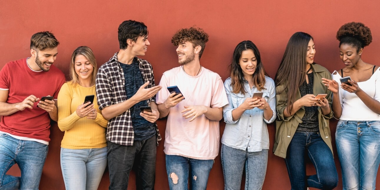 Six young adult friends of different genders and ethnicities stood against a terracotta wall. They are smiling, in conversation and showing each other what's on their phone screens.