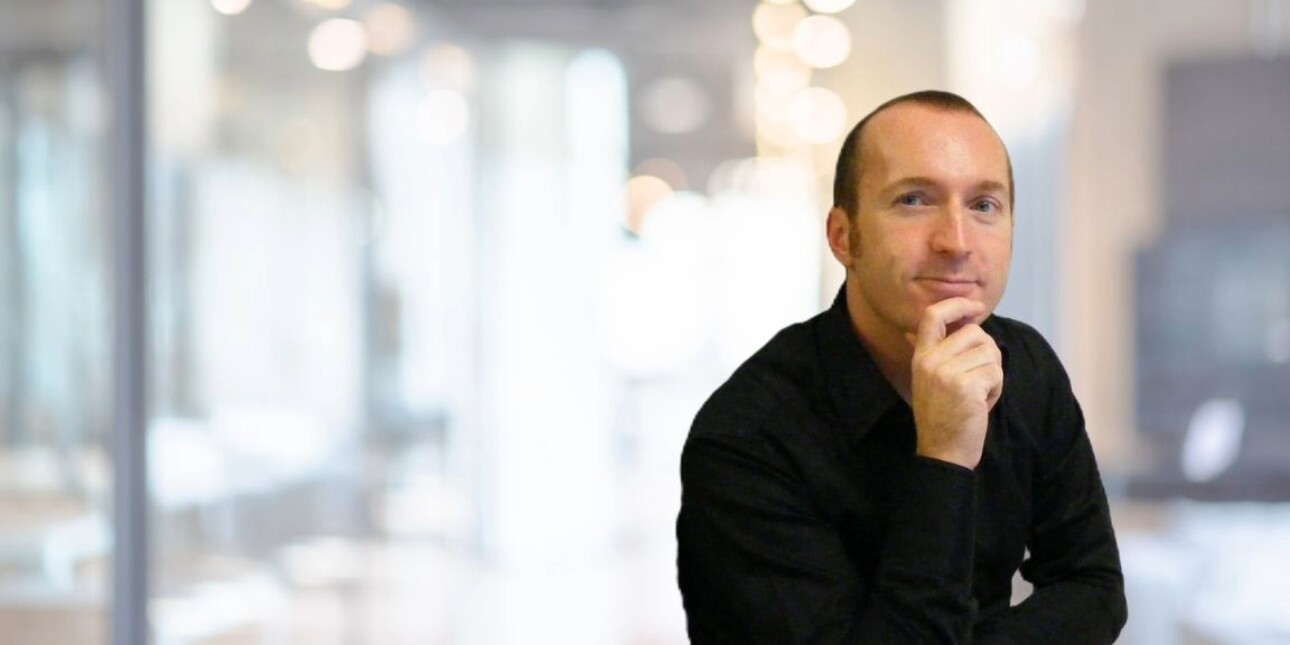 Against the blurred background of an office, Iain Patton sits with his right hand on his chin. Iain is a white man with receding dark hair who wears a black shirt. He is pictured off centre to the right of the image.
