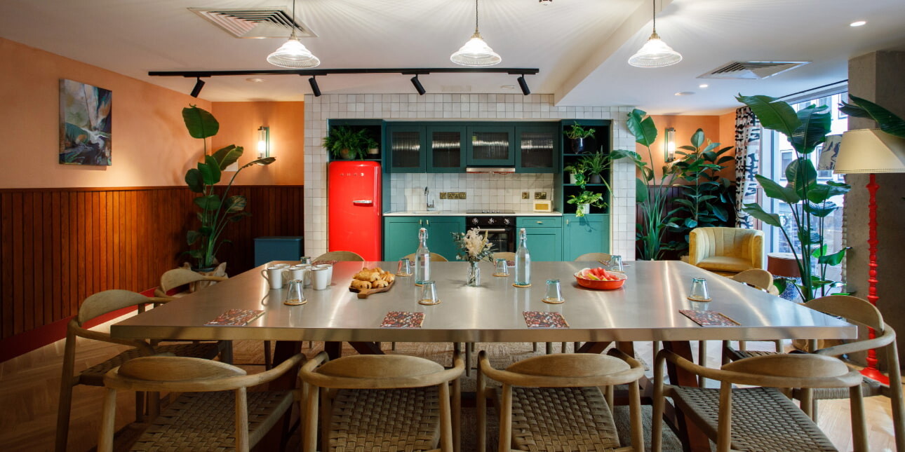 A meeting room at room2 Belfast consisting of a large light coloured table and 12 chairs, in front of a turquoise kitchen with red fridge.