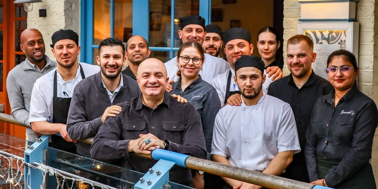 Thirteen people stood looking at the camera on a balcony outside a blue framed door. They mostly wear chefs whites or dark waiters' uniforms.