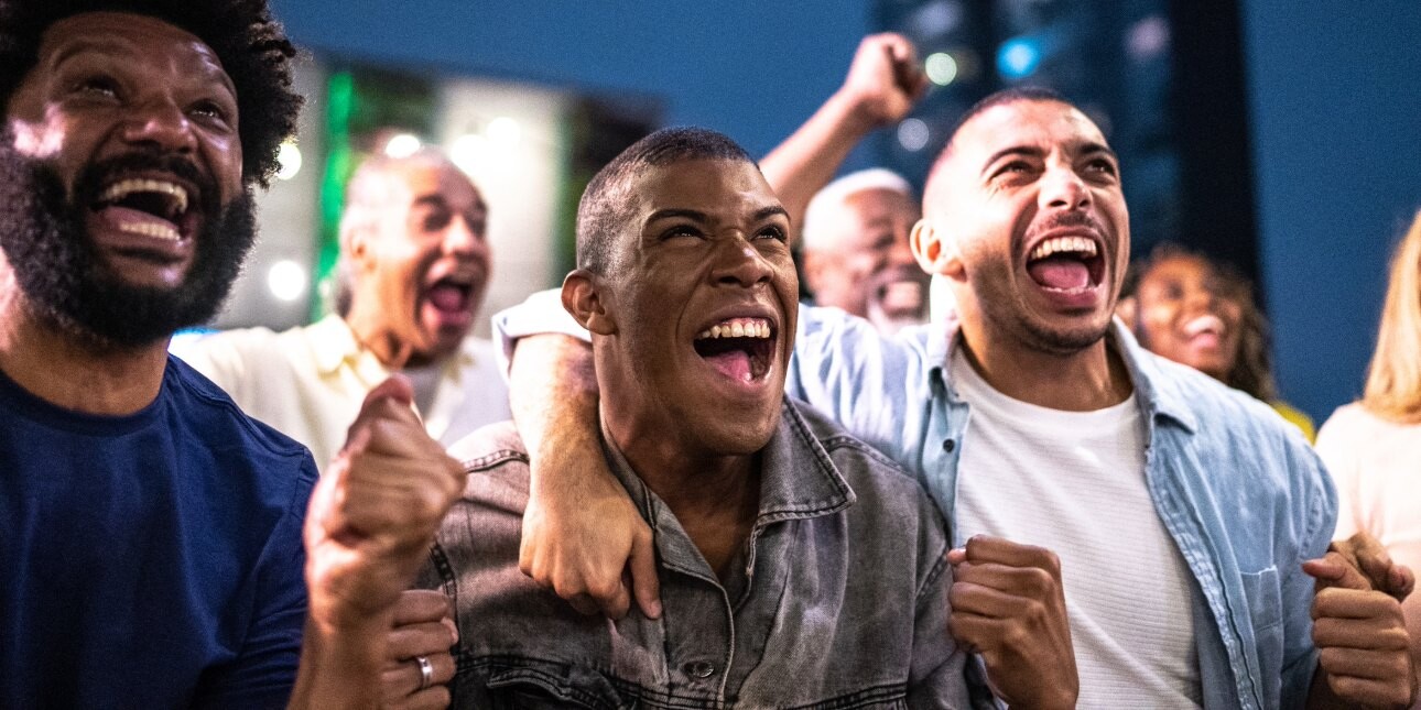 Three male friends cheering and celebrating likely sports action off-camera. Other people perform similar gestures in the background.
