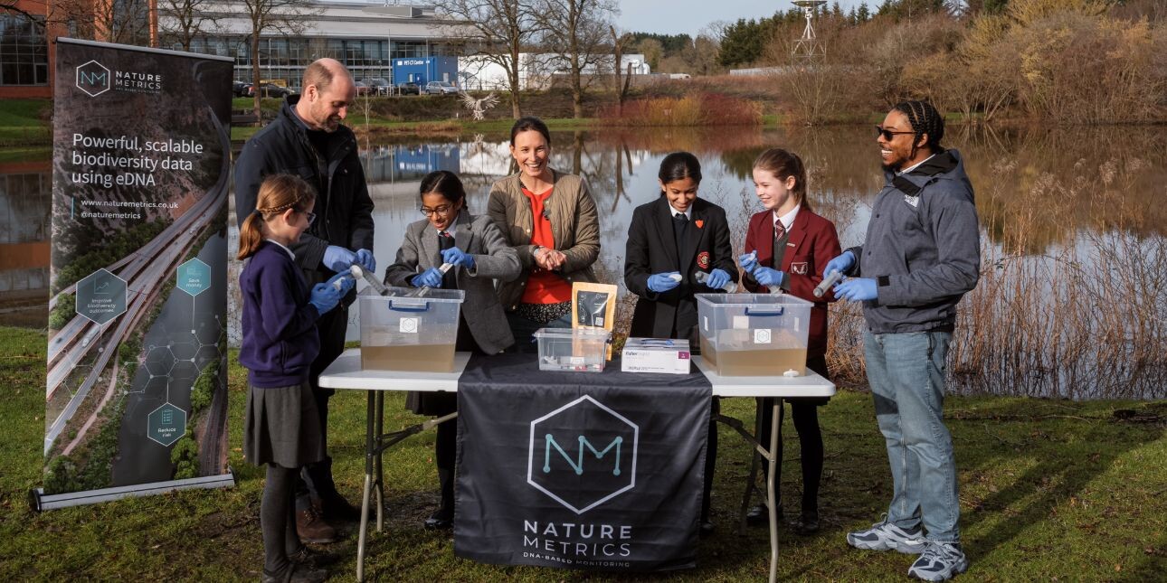 On a patch of grass in front of a pond, Prince William, Dr Kat Bruce and Big Manny stand smiling as four schoolchildren load large syringes with pond water.
