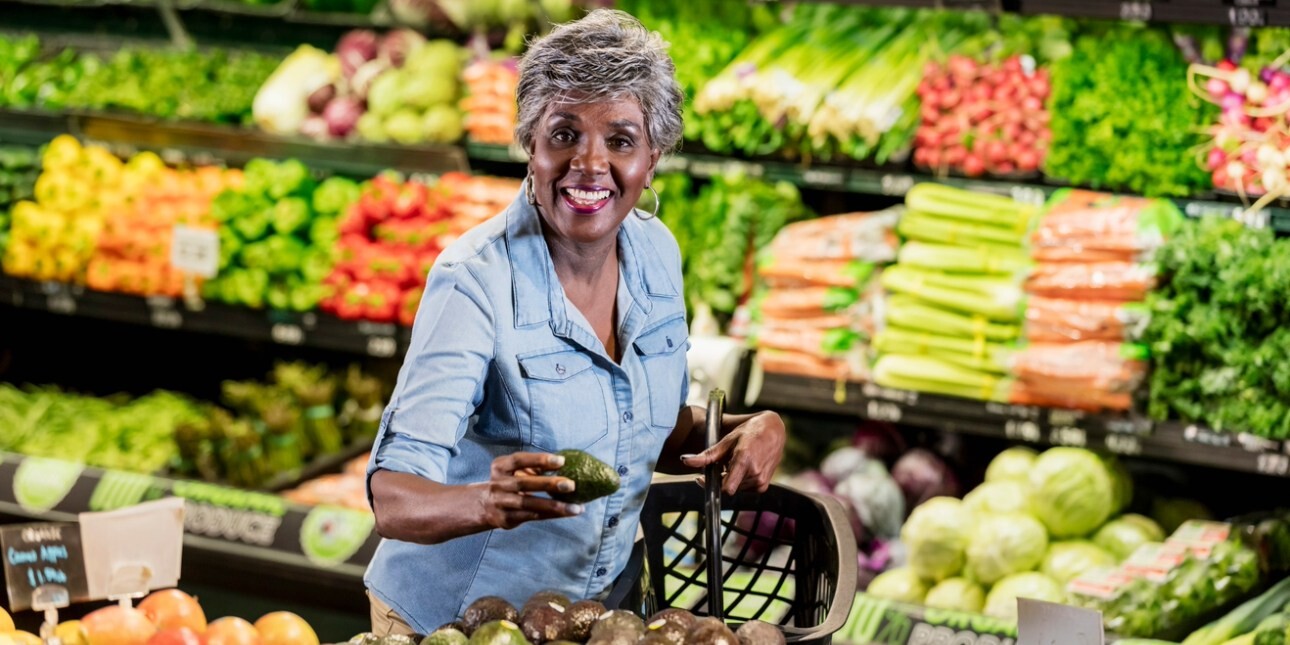 In a supermarket aisle surrounded by displays of fruit and vegetables a mature, smiling Black woman with short grey hair reaches for avocados.