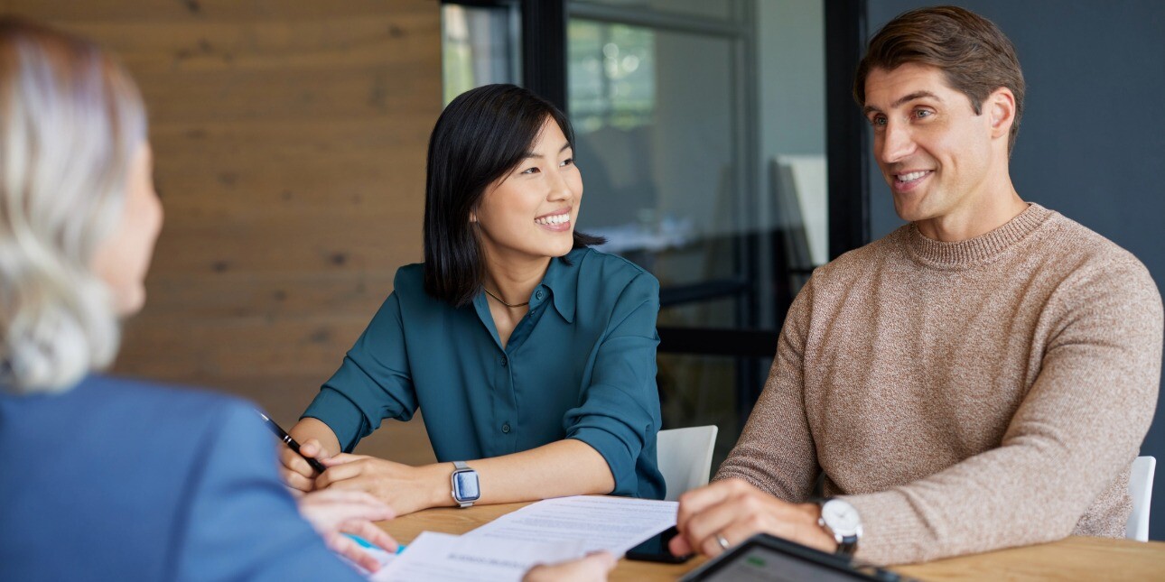 An Asian woman and a white man sit smiling on one side of a board table. Opposite them is the obscured view of another woman
