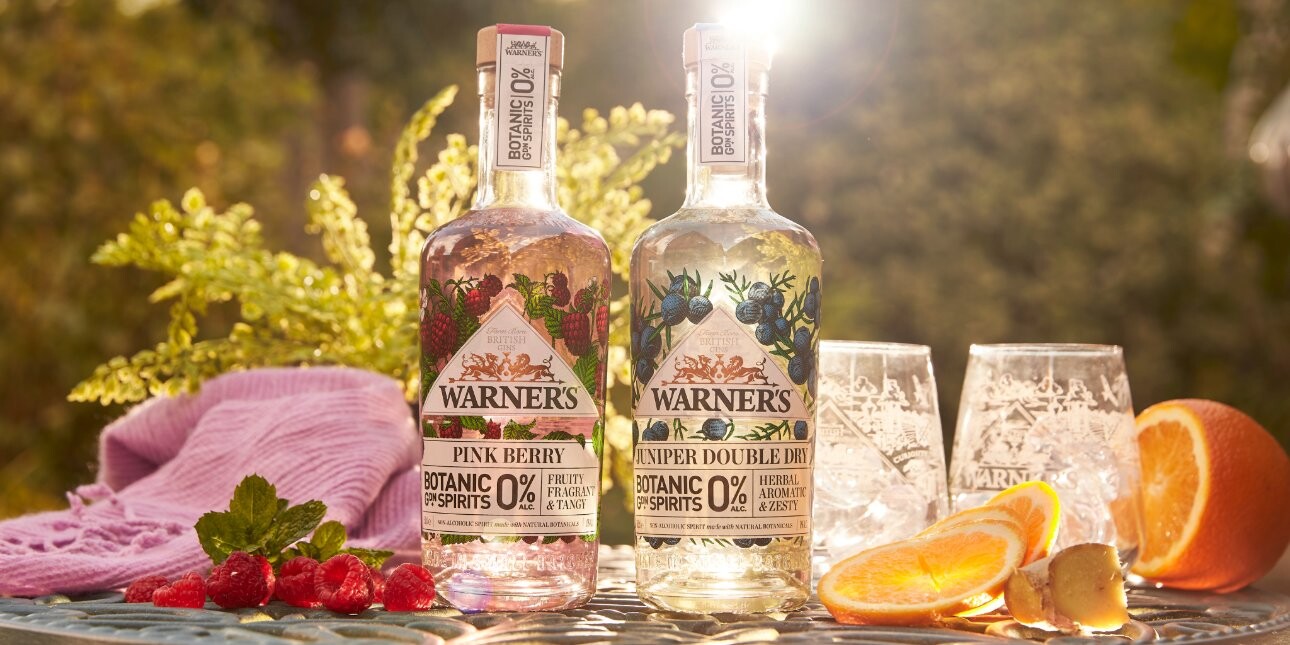 Two bottles of Warners non-alcoholic gin - one clear, one pink - surrounded by glass tumblers, mixed fruit and foliage.