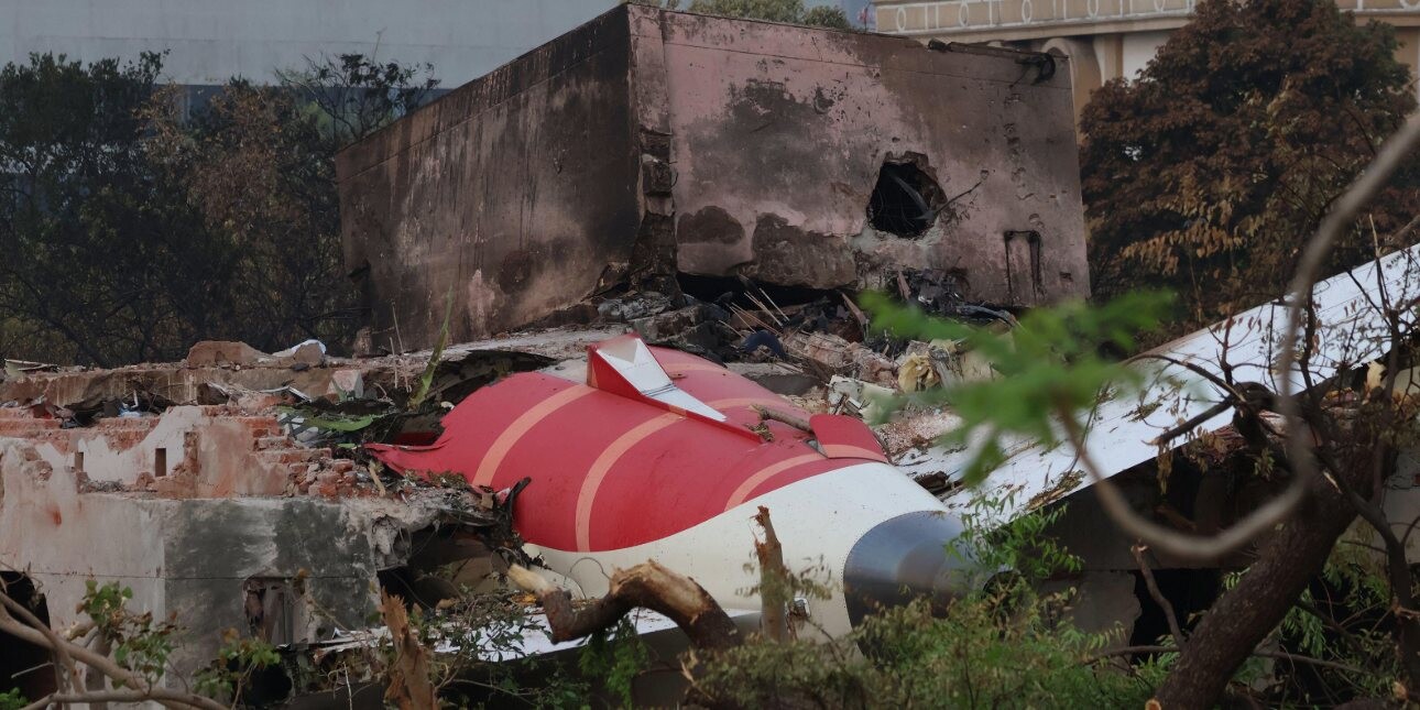 The wrecked red and white tail section of the Air India plane among trees and the side of a building.
