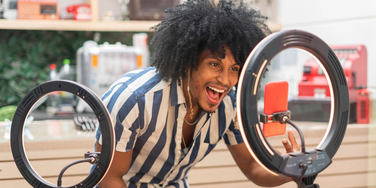 A smiling young Black man wearing blue and white striped shirt while crouching down in front of a mobile phone on a tripod which is flanked by two ring flashes.