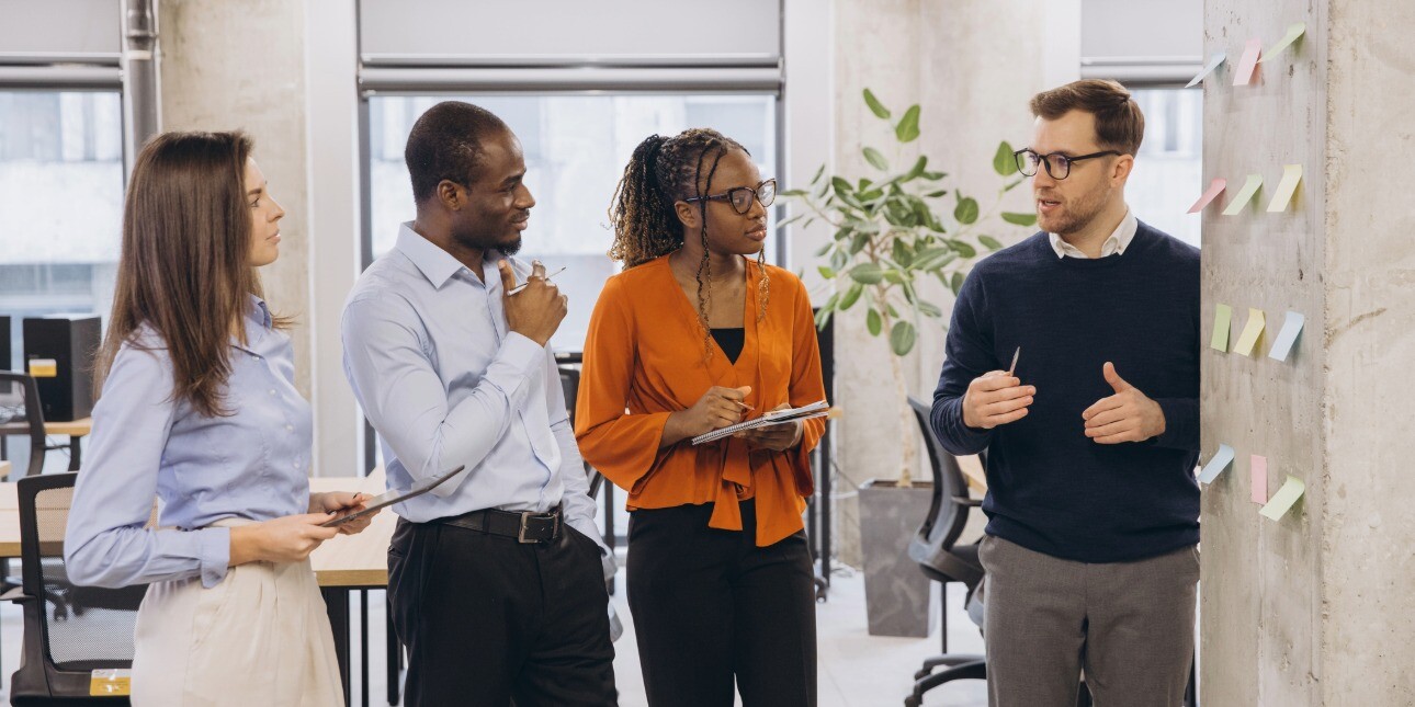 Four colleagues in smart-casual clothing stood in a modern office discussing sticky notes on a cement wall. From left to right: a white woman, a Black man, a Black woman, and a white man.
