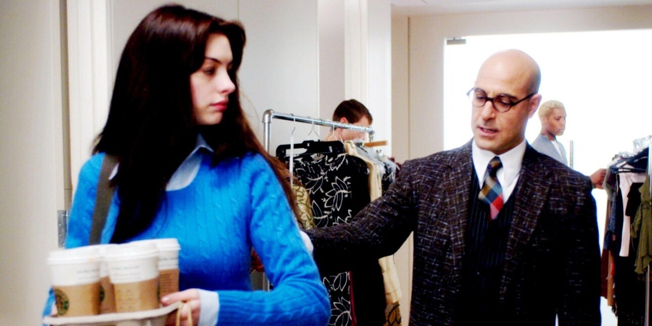A still from the film Devil Wears Prada featuring Anne Hathaway in a blue jumper holding a tray of coffees while Stanley Tucci in a suit is stood behind her.