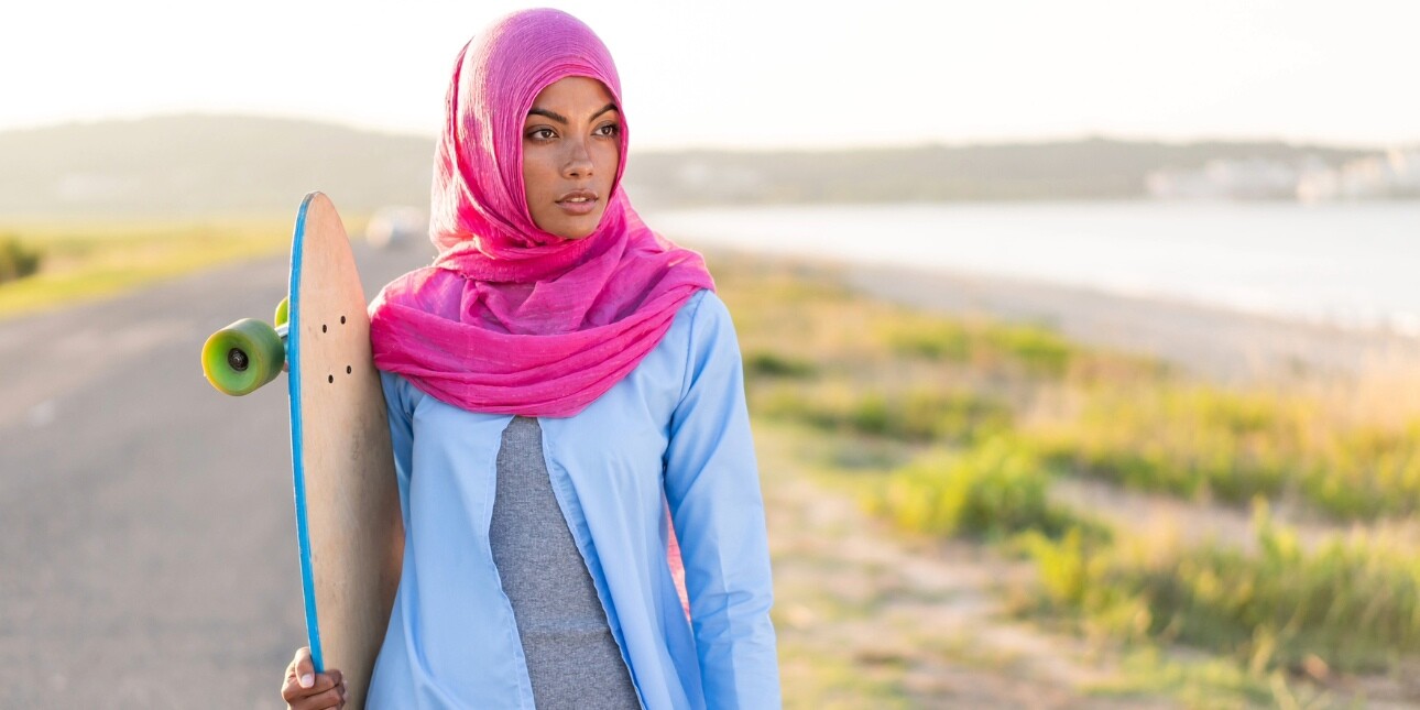 An Asian woman wearing a bright pink hijab and blue abaya over grey clothing, holds a skateboard while stood on an empty road next to grass and an expanse of water