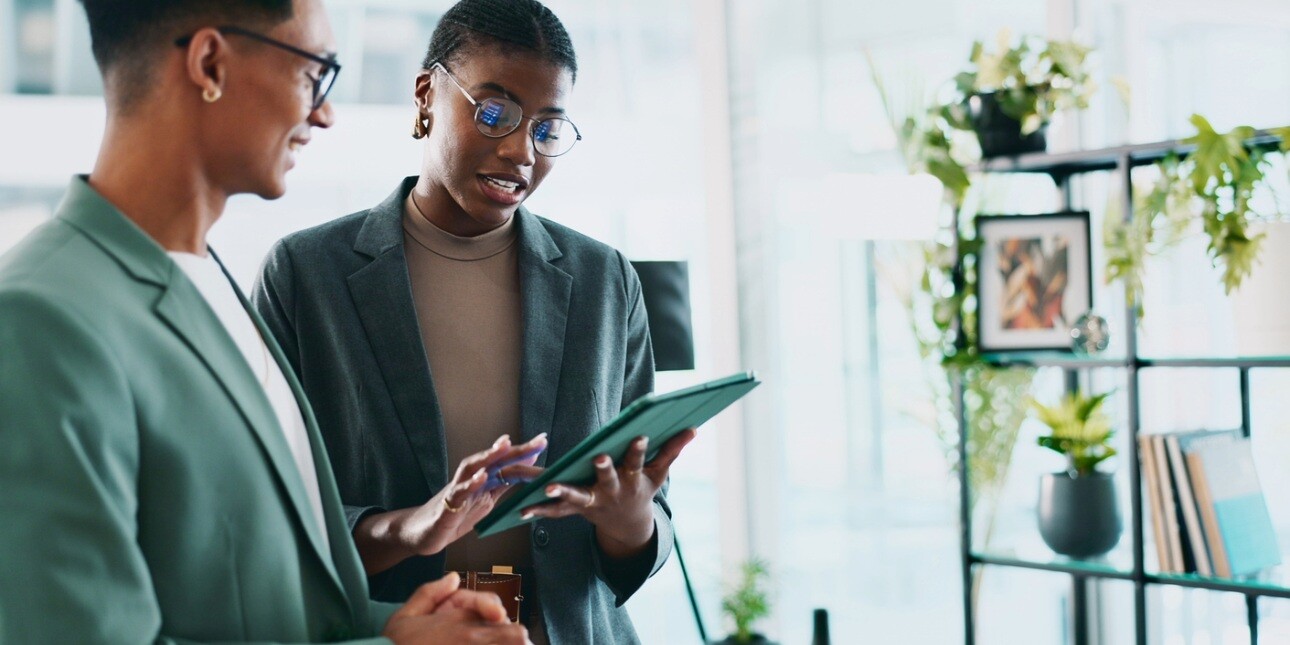 In a modern office two Black male and female colleagues stand looking at a tablet. Both wear dark green smart clothing, hold a green iPad and are surrounded by plants on shelves