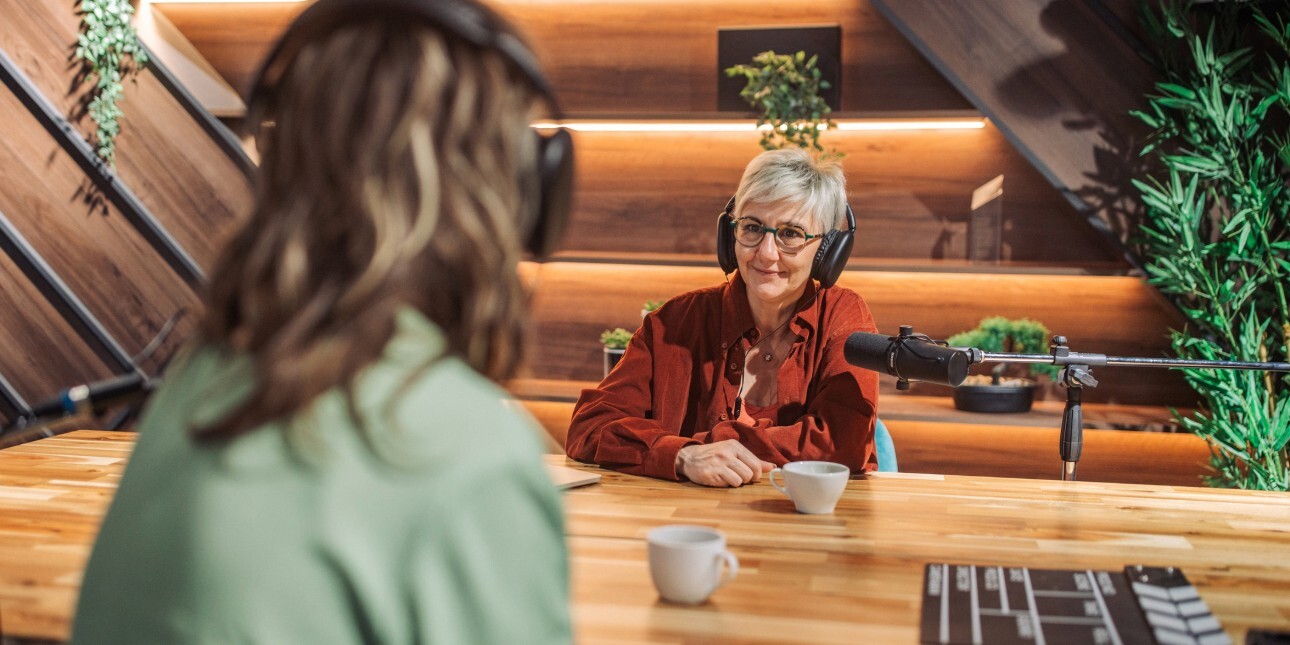 Within the eaves of wood panelled room an older white woman with short grey hair and wearing headphones is in discussion across a microphone with another unidentifiable woman whose back faces the camera