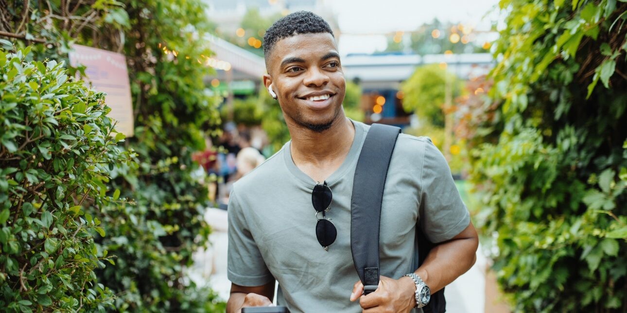 A young smiling Black man wearing earphones and green t-shirts walks in a path surrounded by plants