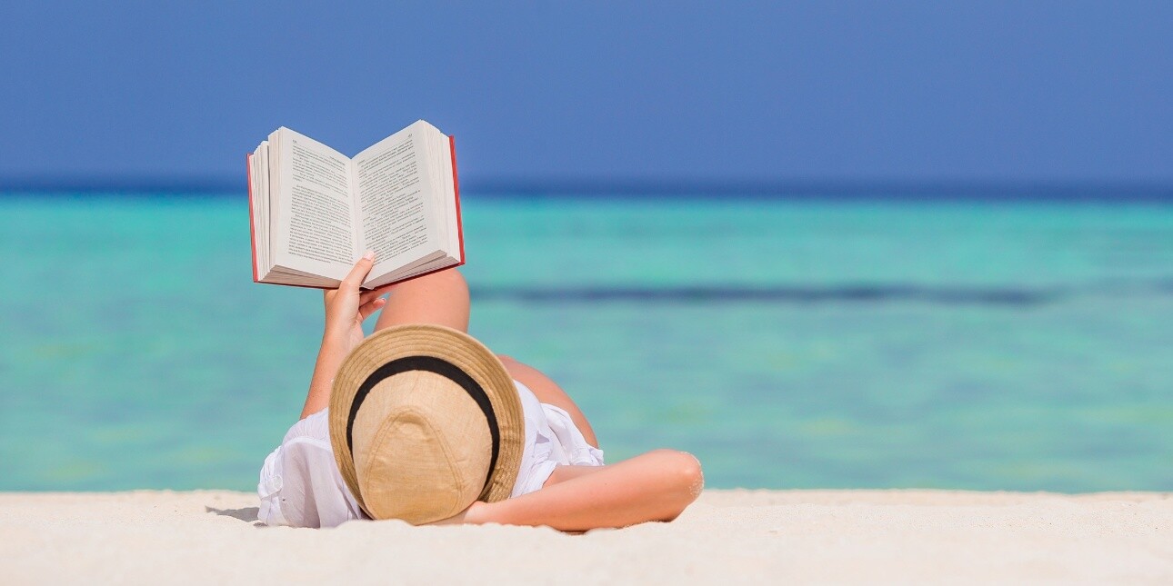 A low angle shot of a person wearing a hat lying on a sandy beach holding a book above them. Behind them are a two-tone blue sea and sky.