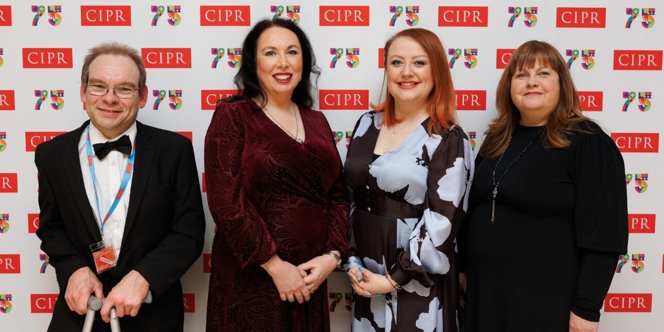 A white man and three white women wearing formal attire, stood smiling in front of a display board with the CIPR 75 logos.