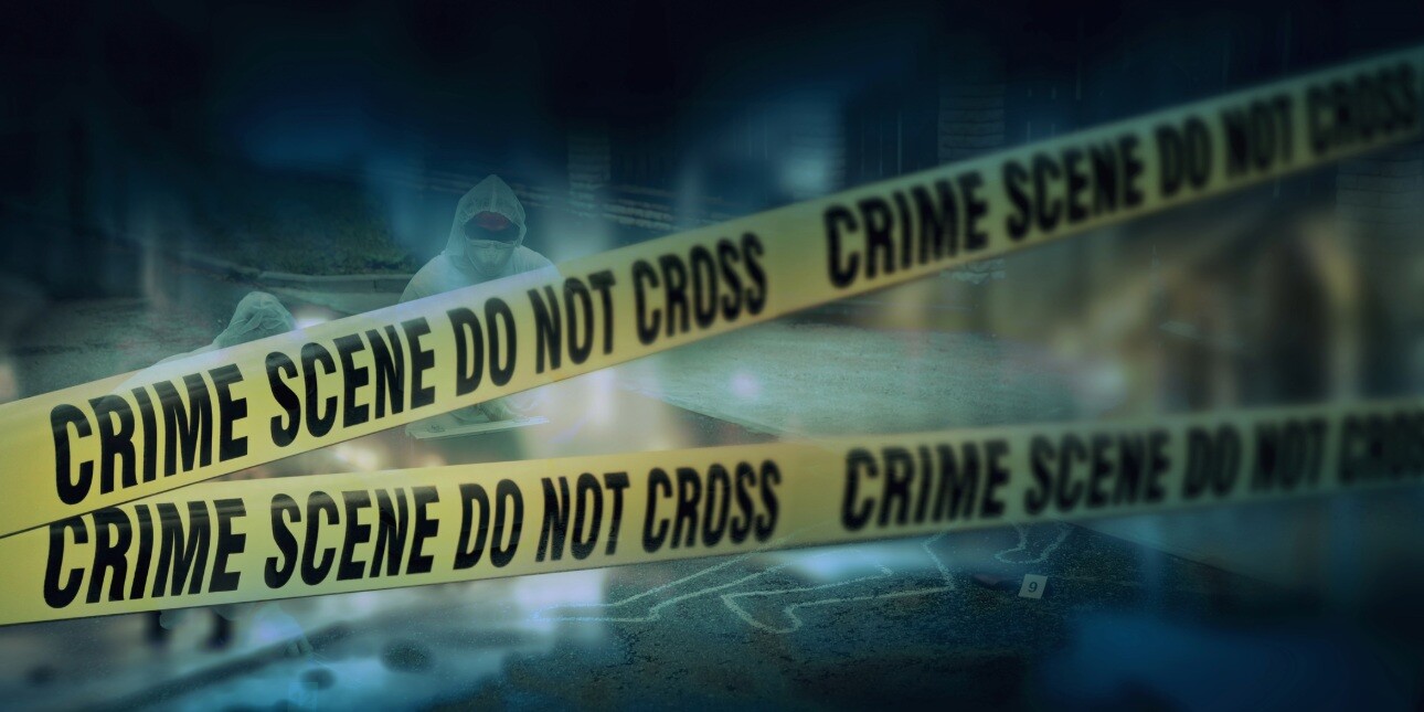 A dark photo with two lines of criss-crossed police tape in the foreground. Behind are two investigators in white protective clothing examining the ground where a white outline of a body can be seen