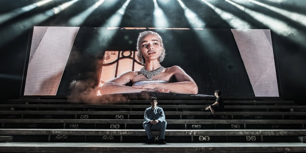 Rachel Zegler leans over a balcony on a large screen at the back of a stage. In front of her on steps sit two men.
