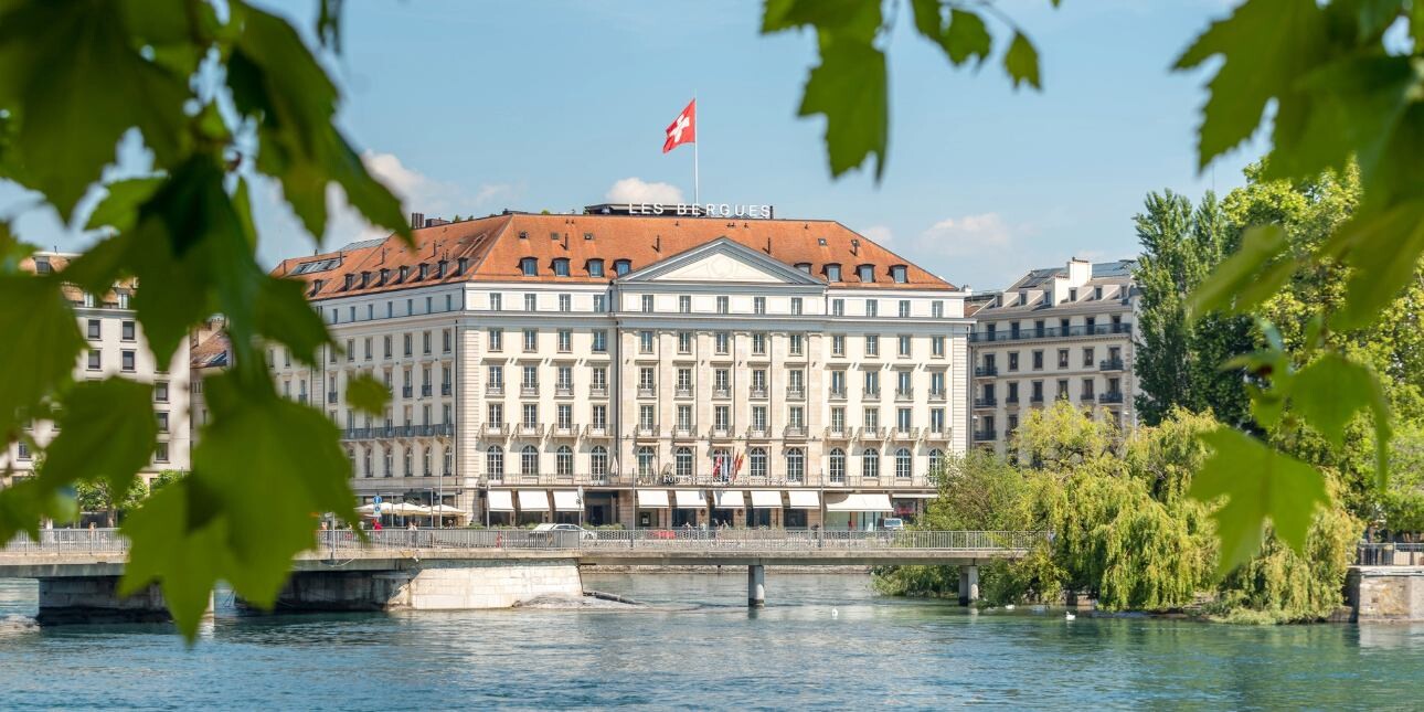 Photographed through the trees, the neoclassical exterior of the Four Seasons in Geneva. In the foreground is the blue water of Lake Geneva, a bridge and leaves. The building imposing five floor facade is topped with a red roof and Swiss flag flying