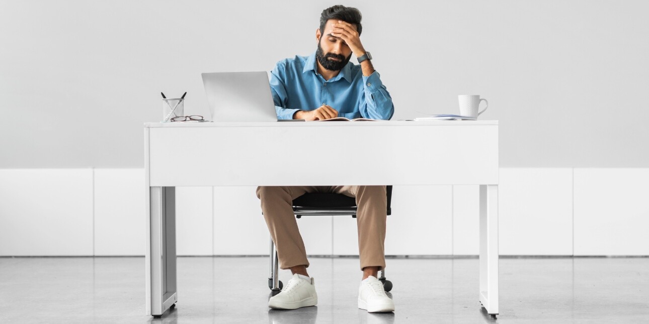 A stressed Asian man with dark hair and beard and wearing a blue shirt and sand beige chinos, sits behind a desk holding his head in his hand. The surroundings are modern with white units and polished concrete floor