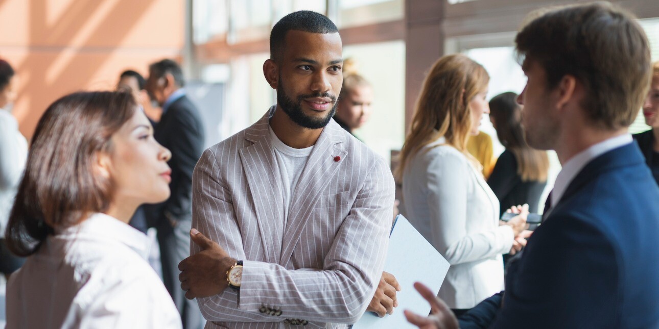 People stood conversing at a busy networking event. In the foreground an Asian woman wearing a white shirt, a Black man in a grey jacket, and a white man in a navy jacket chat to one another.