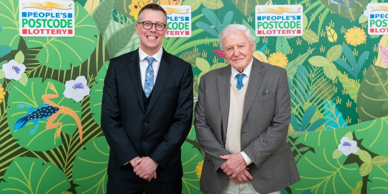 David Miller and Sir David Attenborough stood against a background composed of illustrated leaves and fauna. Both are white men with short hair wearing suits.