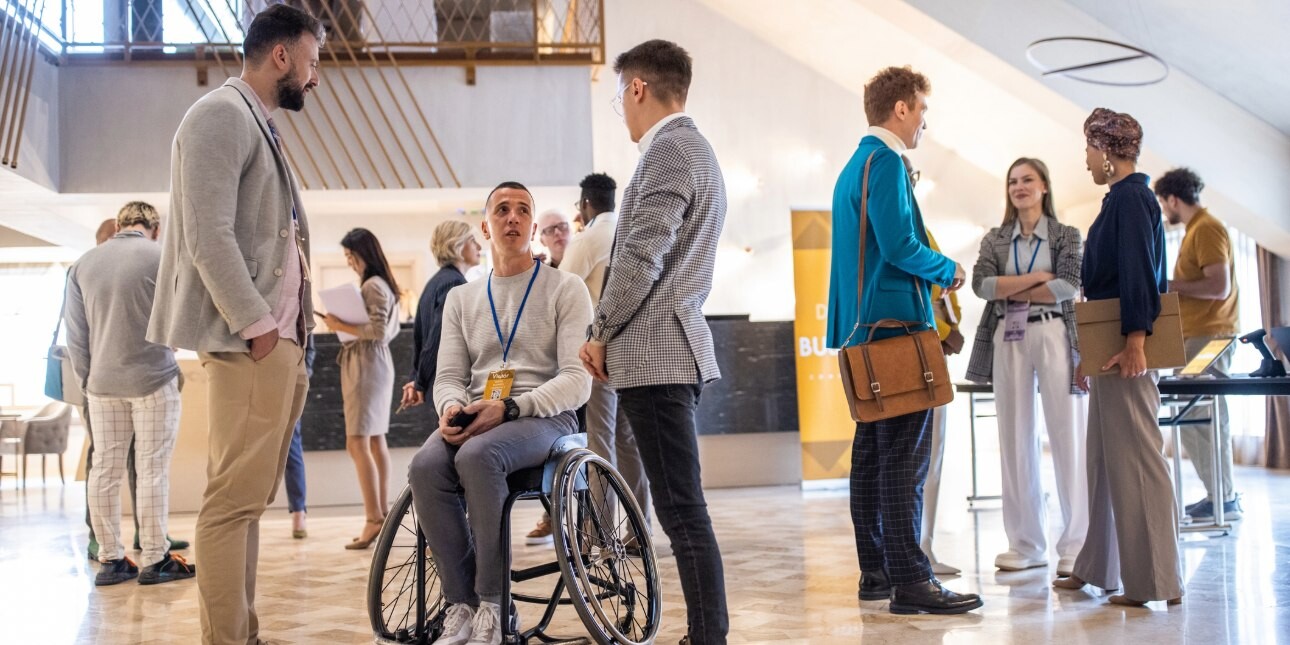 Three men, dressed in smart-casual clothes, chat in a group among a crowd at a light-filled conference reception hall. The man in the middle uses a wheelchair. Other men and women can be seen in the background.