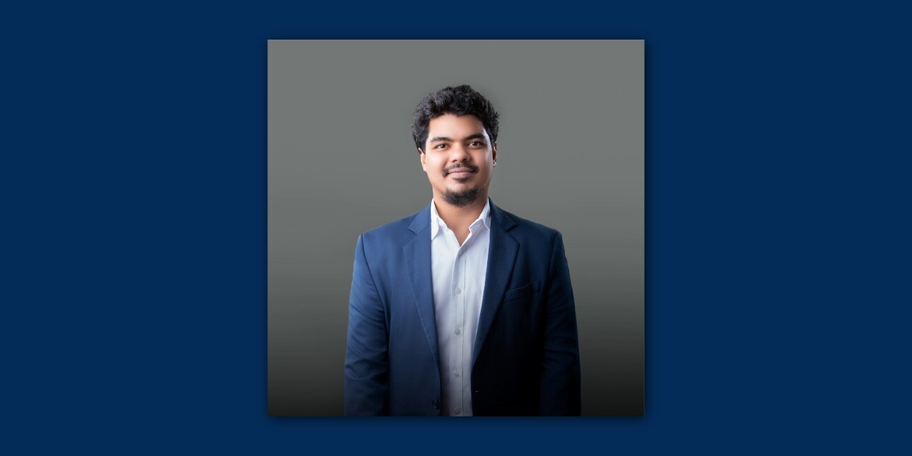 On a dark navy background a colour portrait of Thanzyl Thajudeen wearing a light blue shirt and navy suit. Thajudeen is an Asian man with short dark hair who smiles at the camera.