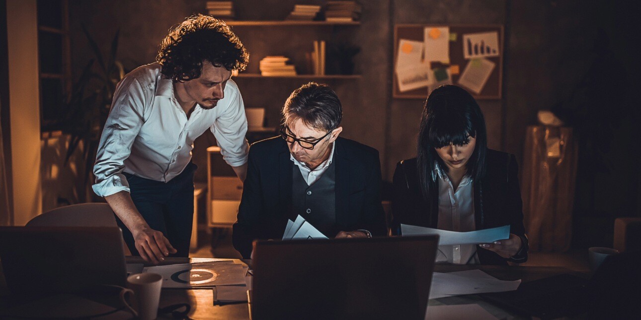 In a dark office, three colleagues work late into the night around a desk.