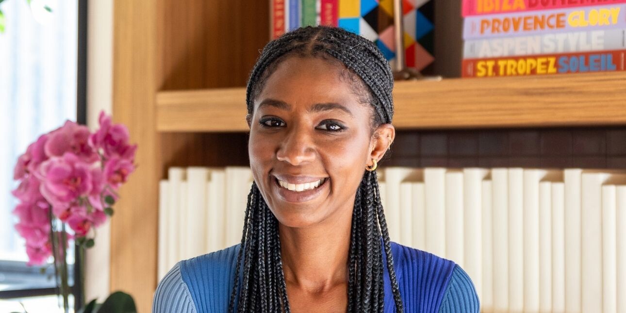 A colour portrait of Terri McCollin smiling. Terri is a Black woman with long braided hair, who wears a two-tone blue top. To her right are pink flowers. There are colourful travel guide behind her.