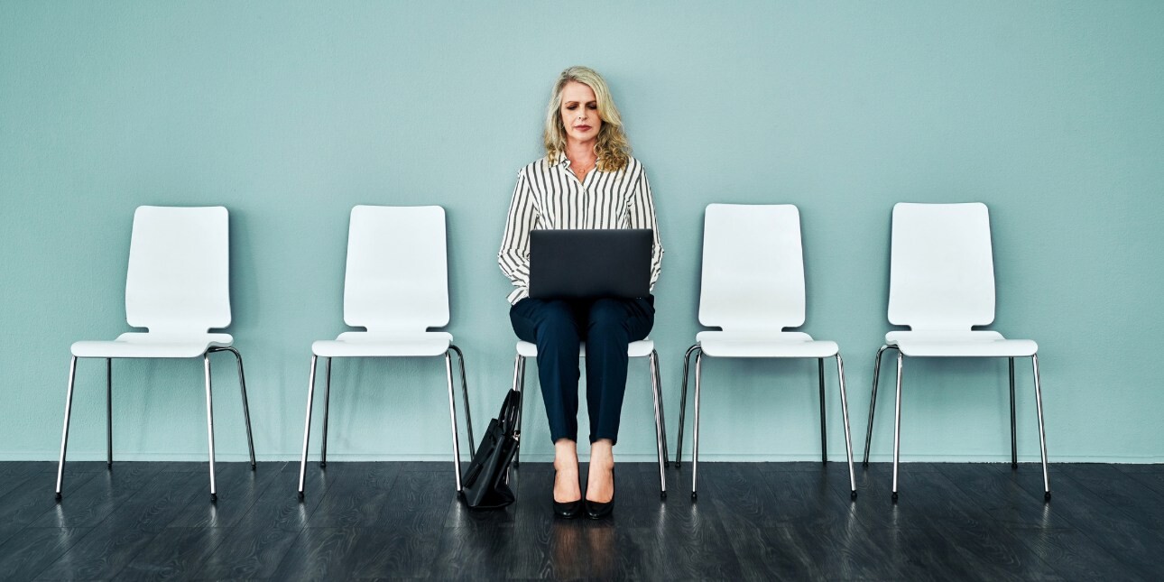 An older woman with blond hair and white top sits in a chair against a turquoise wall looking at the laptop resting on her knees. There are four empty chairs either side of her.