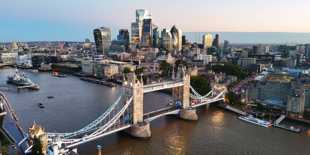 A drone shot of Tower Bridge at dusk. In the background is the Tower of London and dozens of skyscrapers in the City of London.