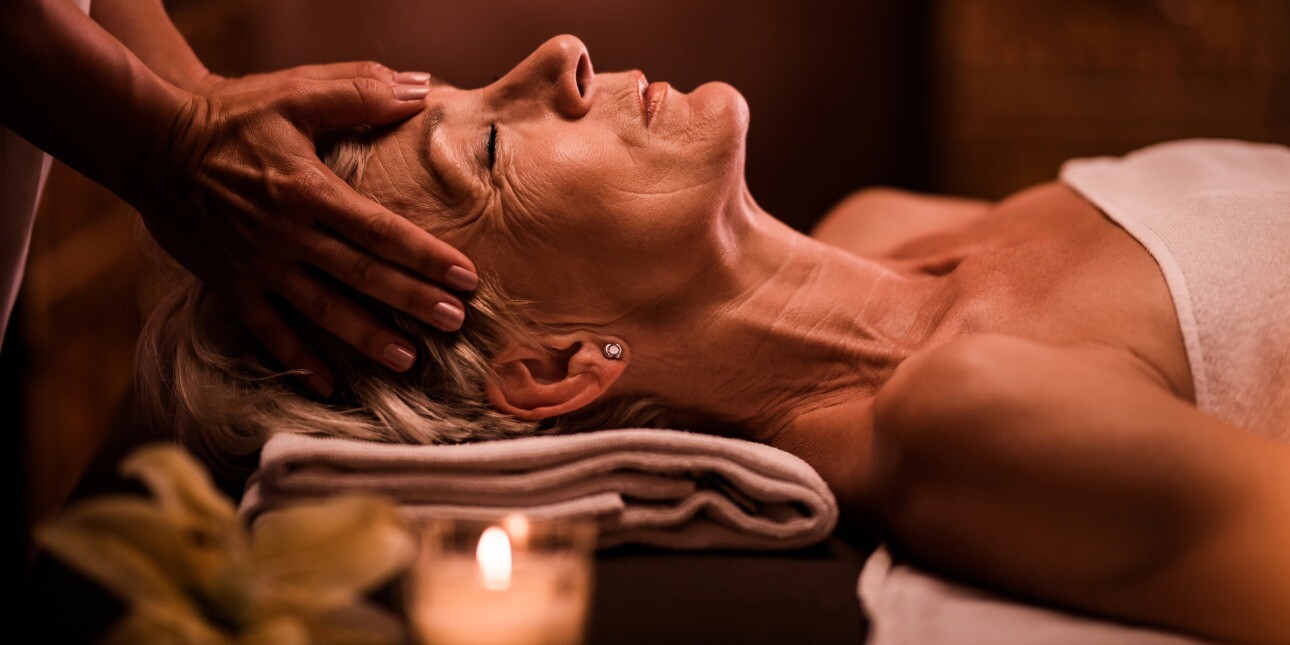 In a dark room lit by candles, a mature white woman with light coloured hair receives a head massage.