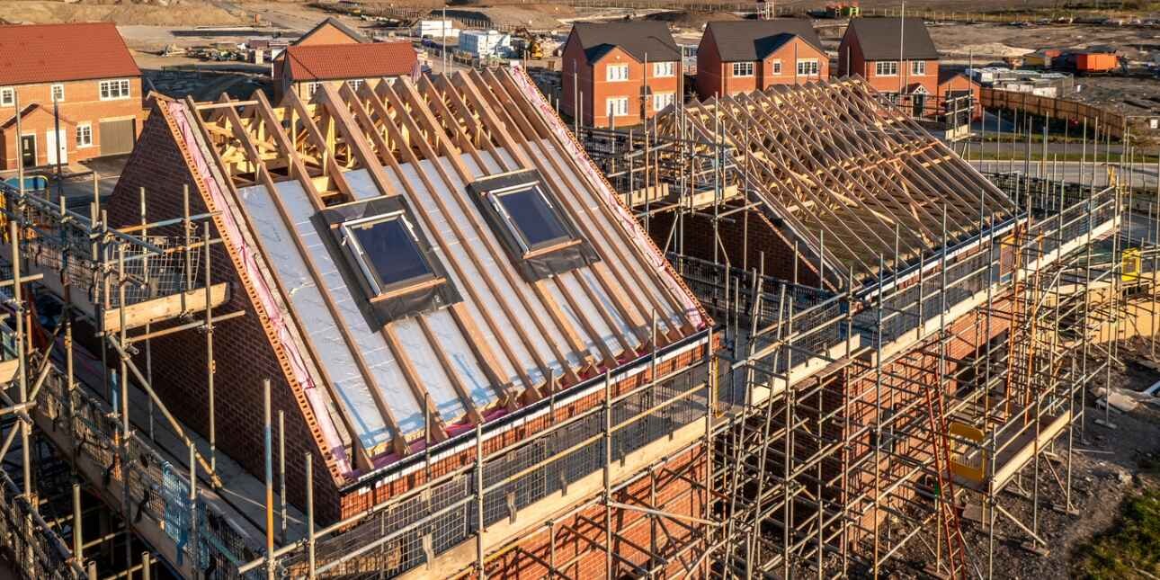 A drone shot of a new home under construction and covered in scaffolding on a new estate, surrounded by other redbrick houses in various stages of completion.