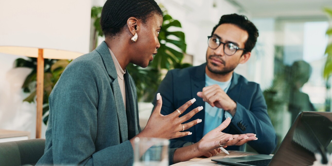 In a modern office environment a Black woman with short hair and wearing a green suit is in discussion with an Asian man with short hair. He wears a navy jacket.