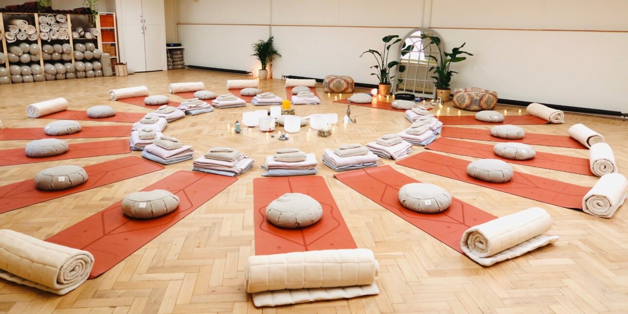 Comfy mats and pillows laid out in a circle on the wood floor of a hall. In the centre of the circle are towels, bowls, lamps and other objects.